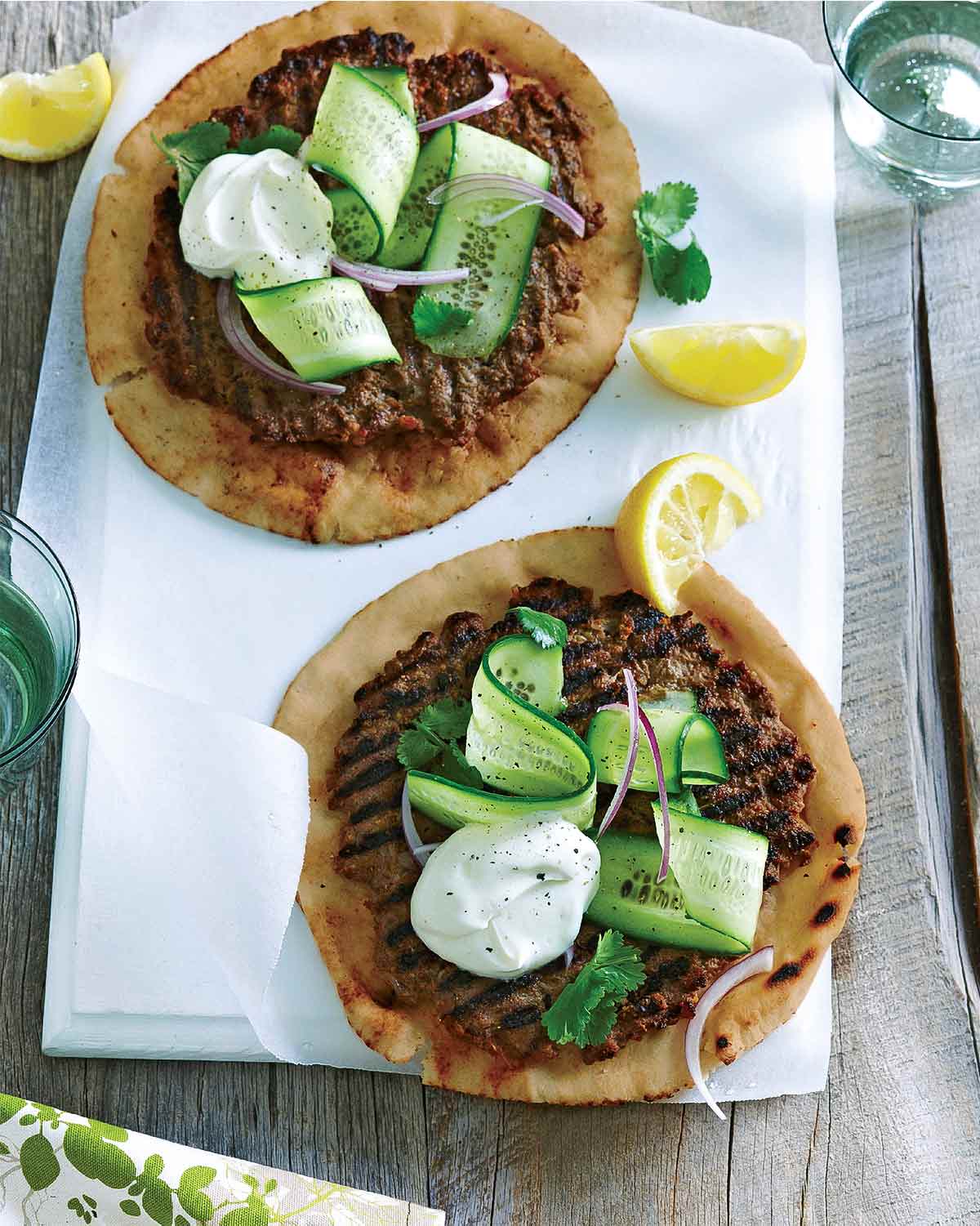 Two servings of spiced beef on whole wheat flatbread topped with cucumber ribbons and yogurt, with lemon wedges on the side.