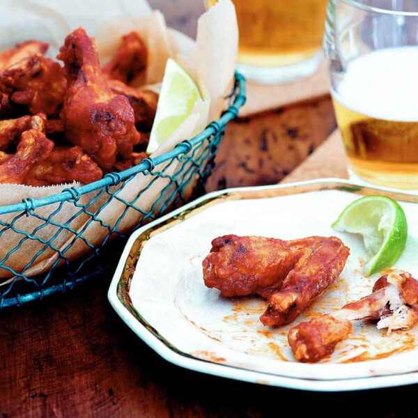 A basket of Sriracha chicken wings, two glasses of beer, and a plate with one eaten and one uneaten chicken wing and a wedge of lime.