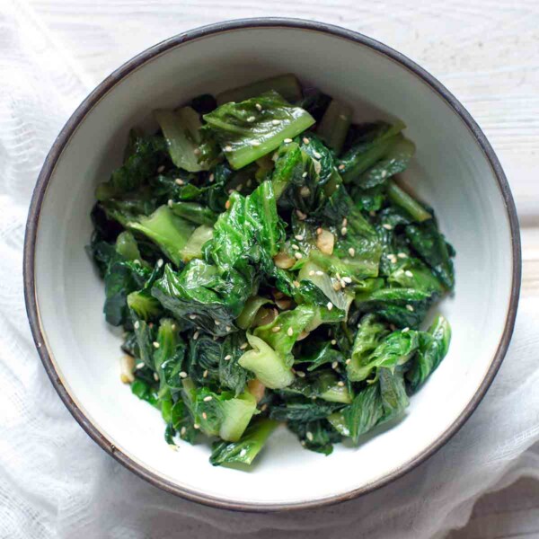 A white bowl filled with stir-fried lettuce, topped with sesame seeds on a white wooden surface.