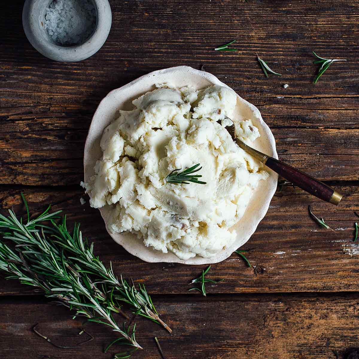 A bowl of vegan mashed potatoes garnished with a sprig of rosemary with a spoon resting inside and more rosemary on the side.