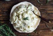 A bowl of vegan mashed potatoes garnished with a sprig of rosemary with a spoon resting inside and more rosemary on the side.