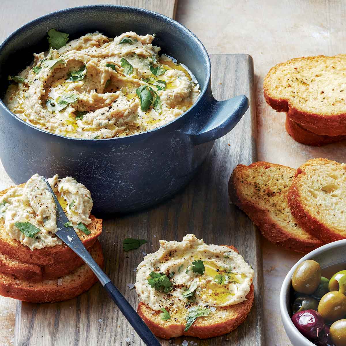 A round blue dish filled with white bean dip, with some spread on crostini scattered around the bowl and a small dish of olives on the side.