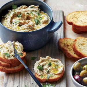 A round blue dish filled with white bean dip, with some spread on crostini scattered around the bowl and a small dish of olives on the side.