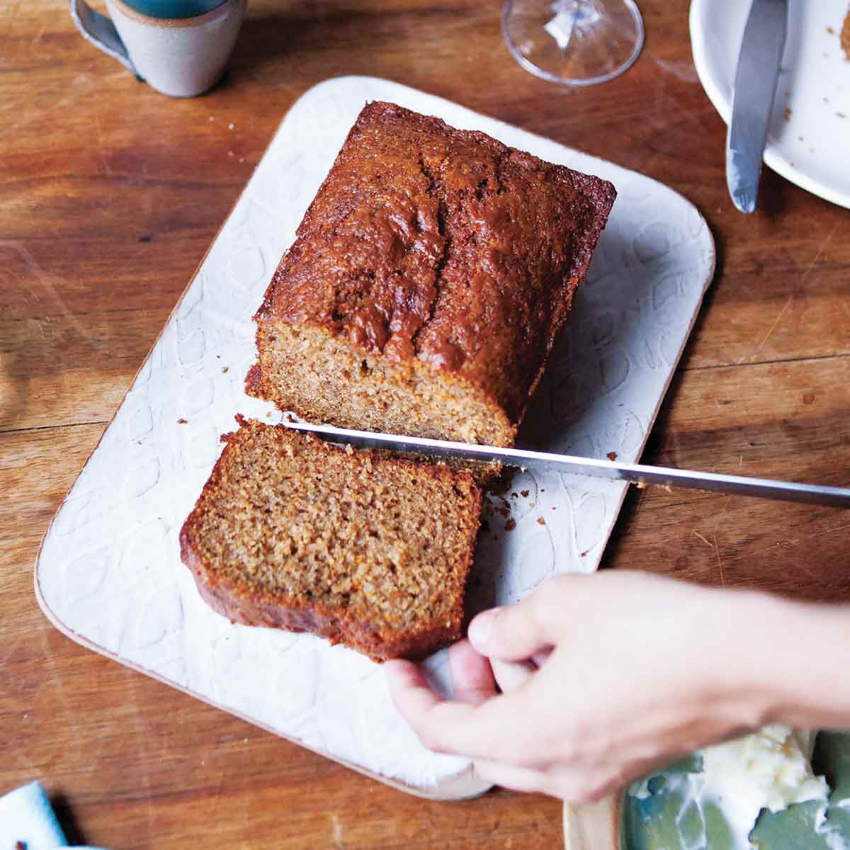 A person cutting a slice from a loaf of whole wheat banana bread on a white cutting board.