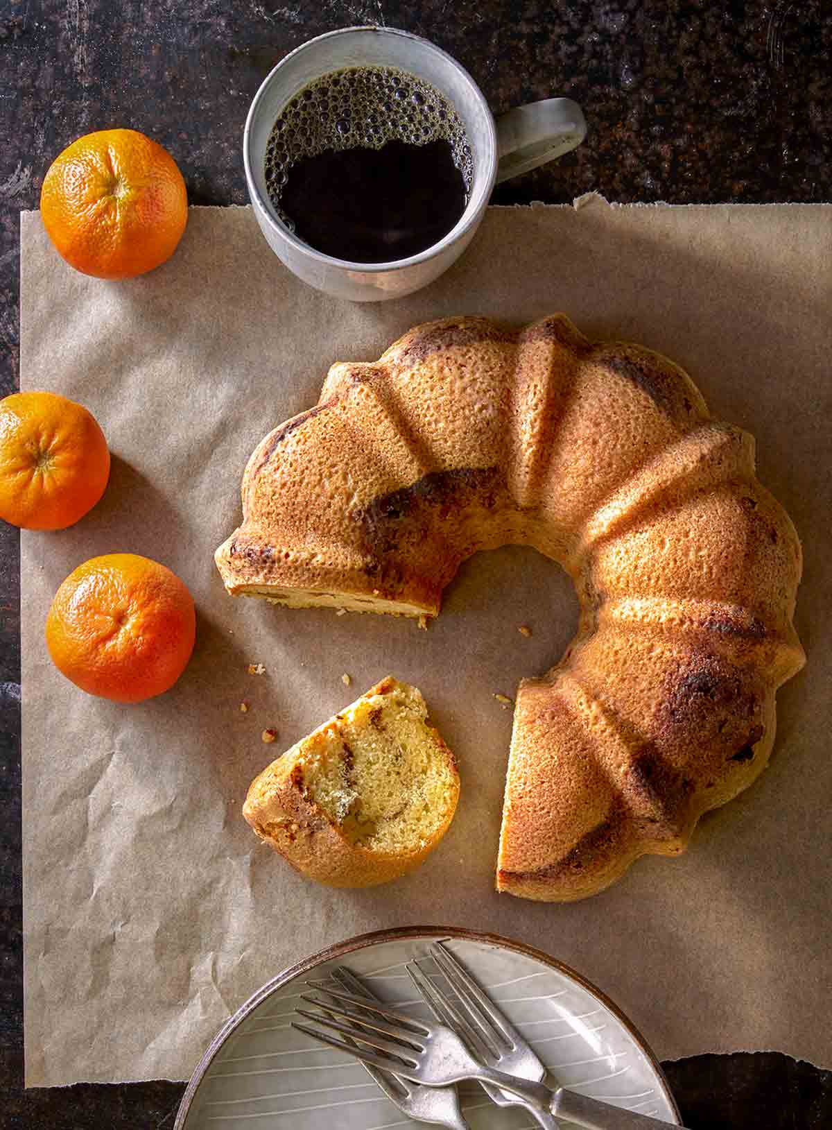 A Zingerman's sour cream coffee cake with a piece cut from it and lying on its side with a cup of coffee, and a stack of plates and pile of forks nearby.