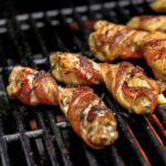 Several bacon-wrapped chicken wings on a grill grate with flame below.