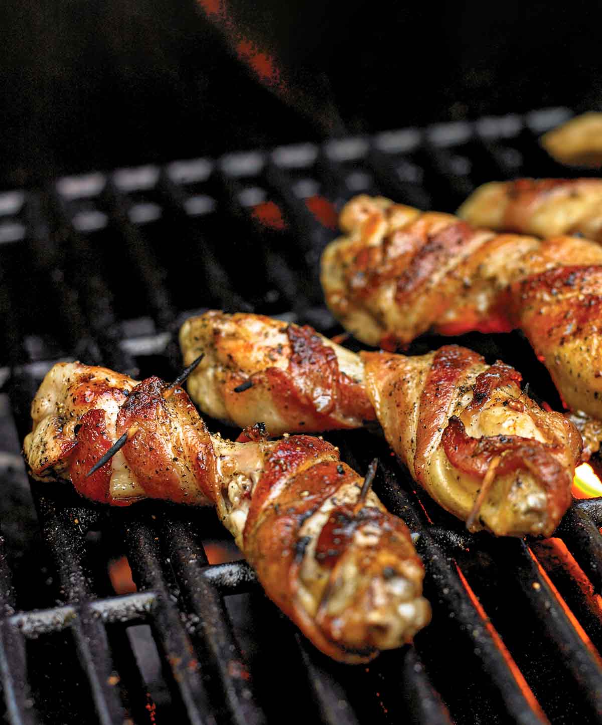 Several bacon-wrapped chicken wings on a grill grate with flame below.