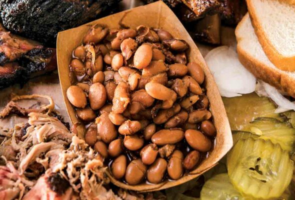 A cardboard food container filled with barbecue beans surrounded by ribs, pickles, and shredded meat.