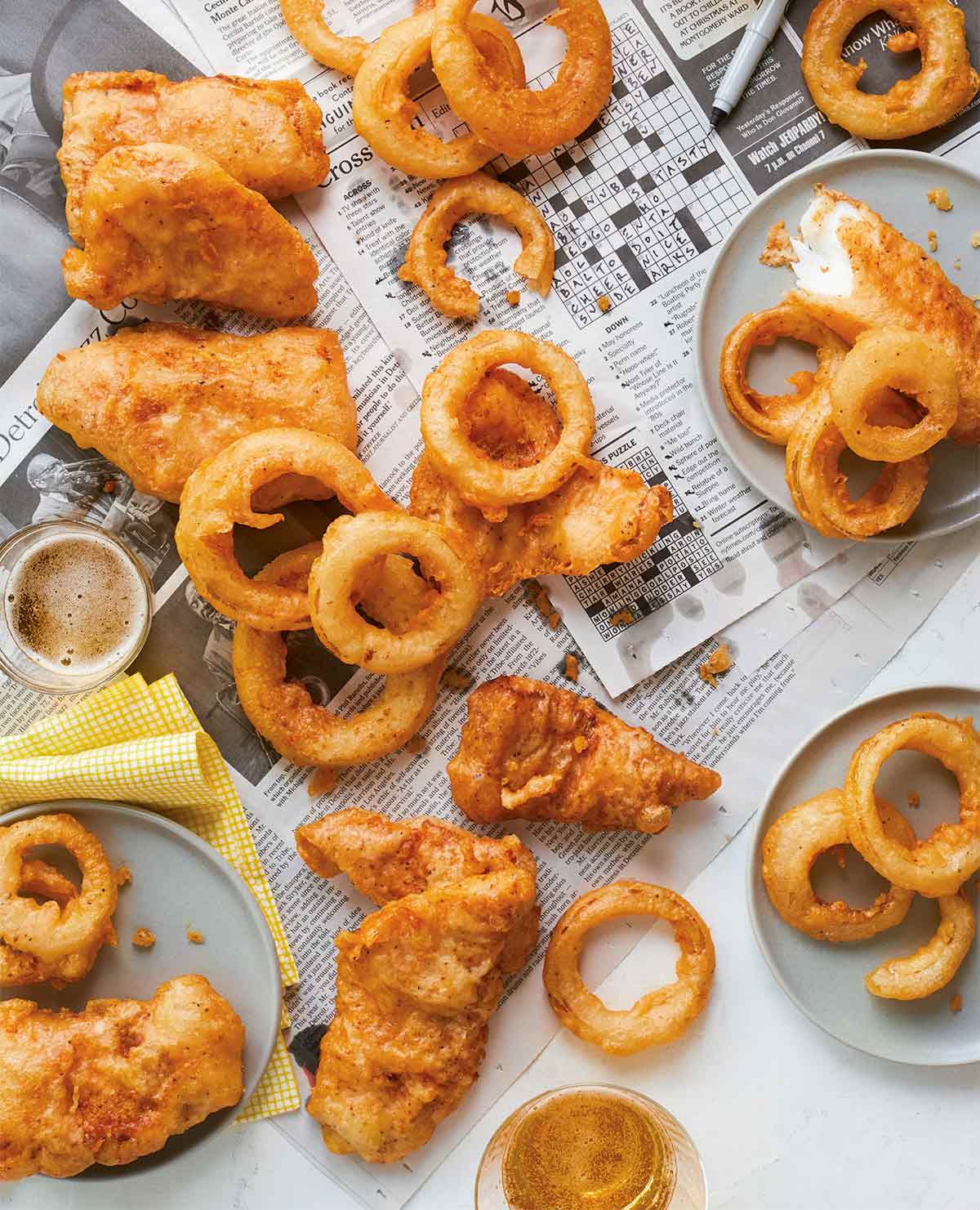 Several pieces of beer-battered fish scattered across newspaper with fried onions rings and two glasses of beer.