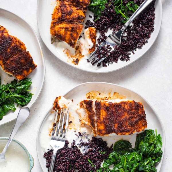 Three white plates with blackened red snapper, black rice, yogurt sauce, and sauteed spinach.