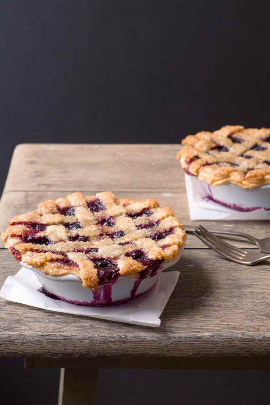 Two individual blueberry pies on on a wooden table with forks beside them.