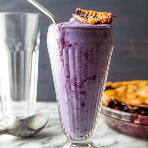 A tall glass filled with blueberry pie milkshake, topped with a piece of pie crust.