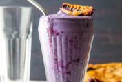 A tall glass filled with blueberry pie milkshake, topped with a piece of pie crust.