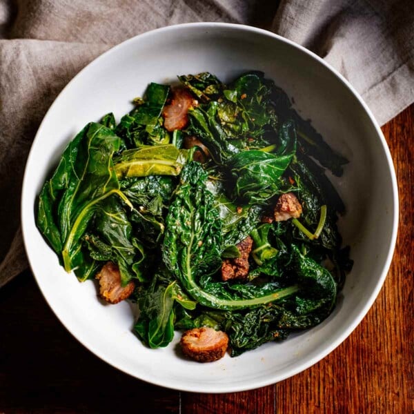 A white bowl filled with braised greens with Andouille sausage on a wooden table with a linen napkin underneath.