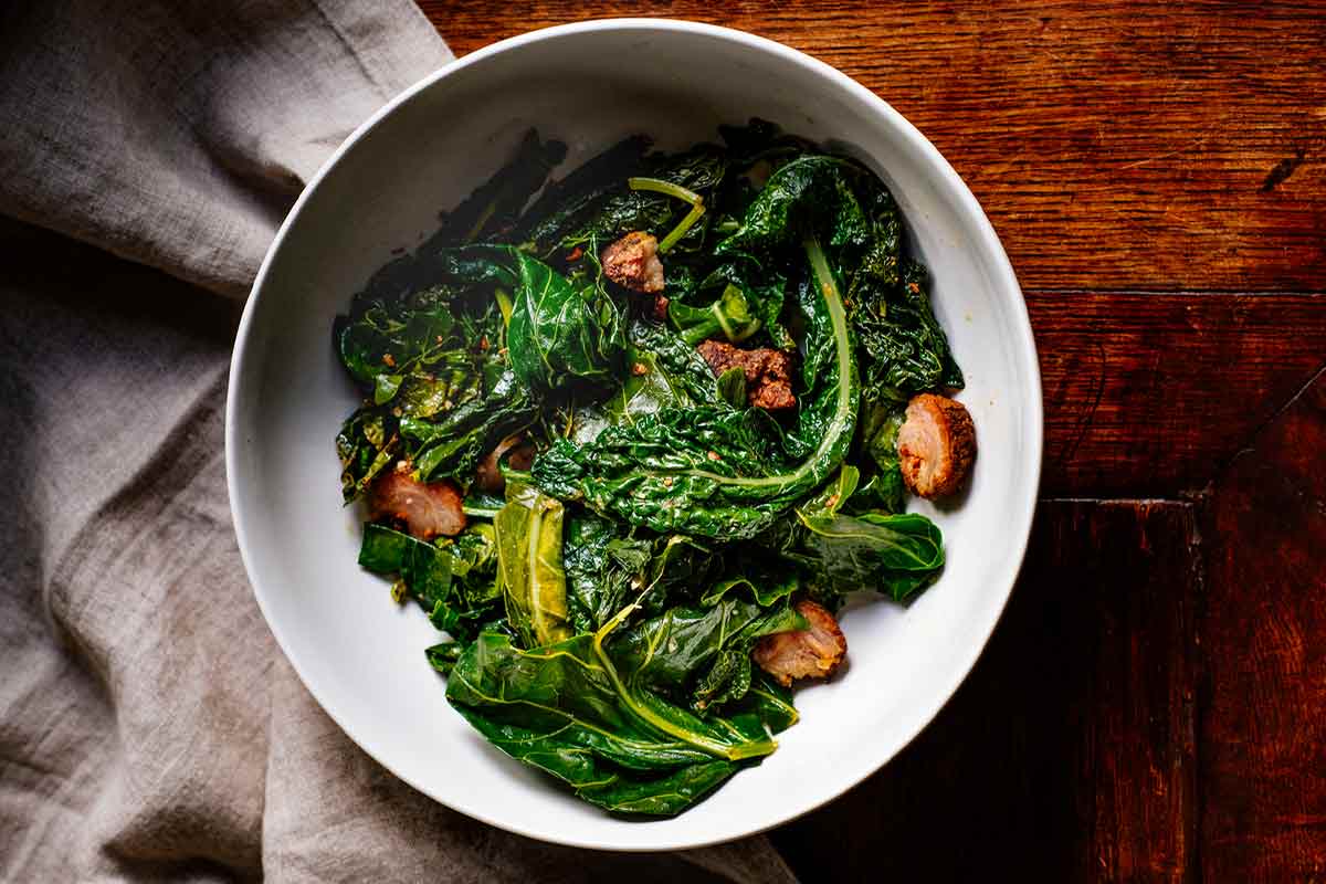 A white bowl filled with braised greens with Andouille sausage on a wooden table with a linen napkin underneath.