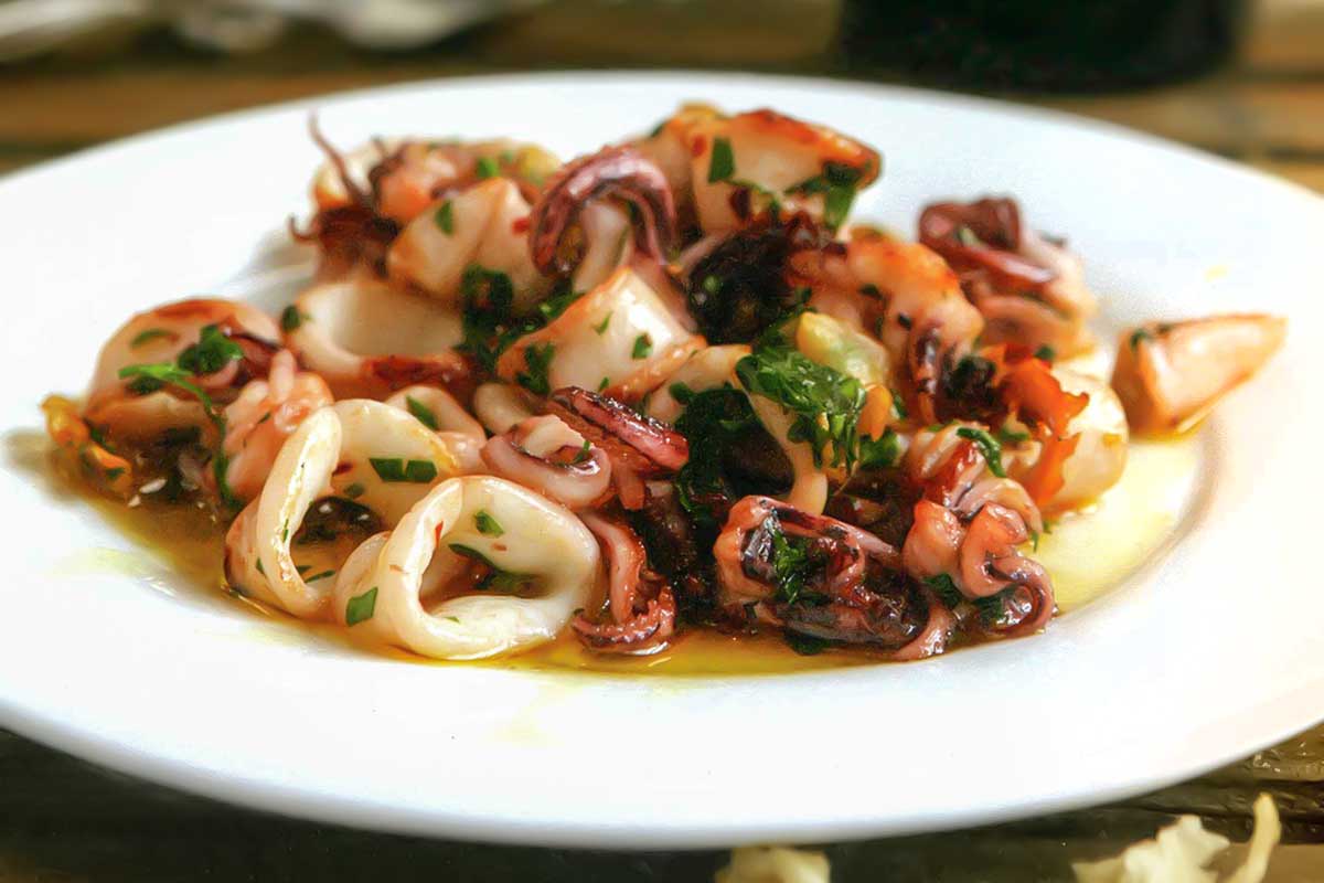 A white plate topped with calamari à la plancha -- seared calamari -- with a bottle of wine in the background.