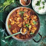 A metal pot filled with cauliflower and tomato curry with a ladle resting inside, and a bowl of rice, a bowl of chutney and a pile of cilantro stems on the side.