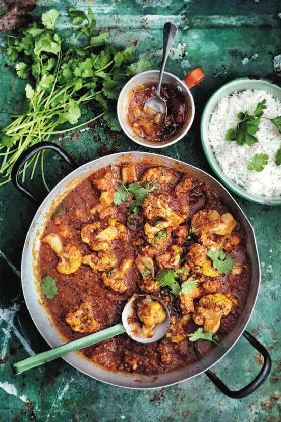 A metal pot filled with cauliflower and tomato curry with a ladle resting inside, and a bowl of rice, a bowl of chutney and a pile of cilantro stems on the side.