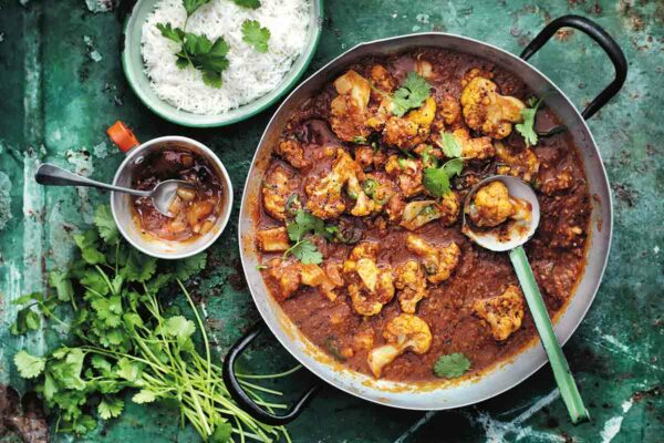 A metal pot filled with cauliflower and tomato curry with a ladle resting inside, and a bowl of rice, a bowl of chutney and a pile of cilantro stems on the side.