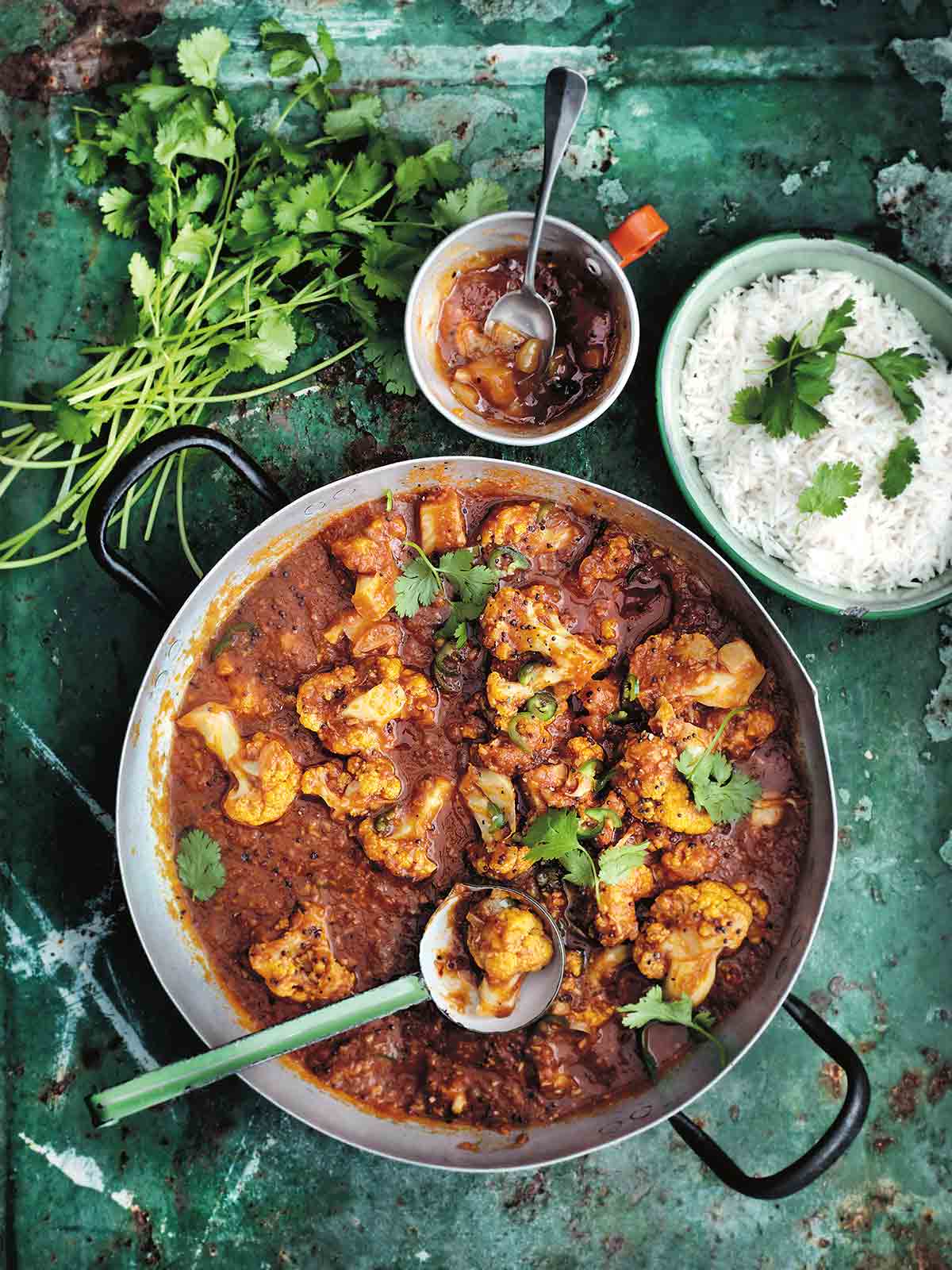 A metal pot filled with cauliflower and tomato curry with a ladle resting inside, and a bowl of rice, a bowl of chutney and a pile of cilantro stems on the side.