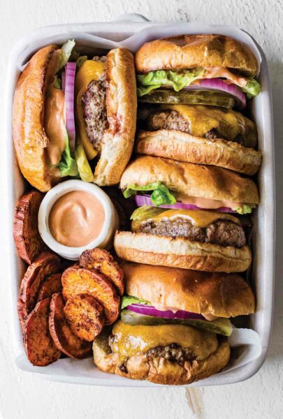 Four cheeseburgers with everything sauce tucked into a white casserole dish with sweet potato chips and a dish of everything sauce.