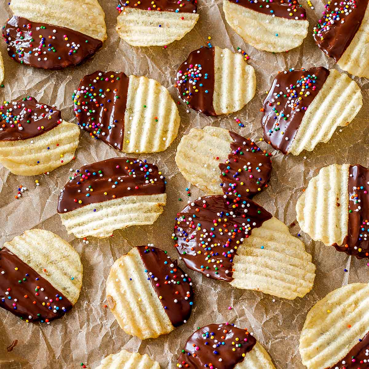 Chocolate covered potato chips with sprinkles on the chocolate in a single layer on a parchment-lined baking sheet.