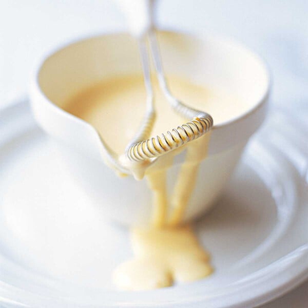 A dish of classic Hollandaise sauce on a white plate with a whisk resting on the dish.