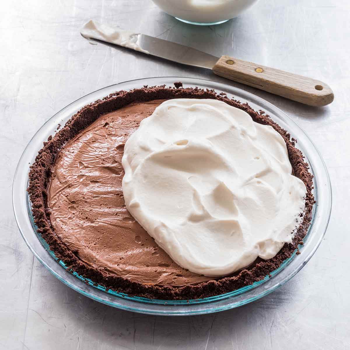 A dark chocolate cream pie partially covered with whipped cream and a knife and a bowl of whipped cream on the side.
