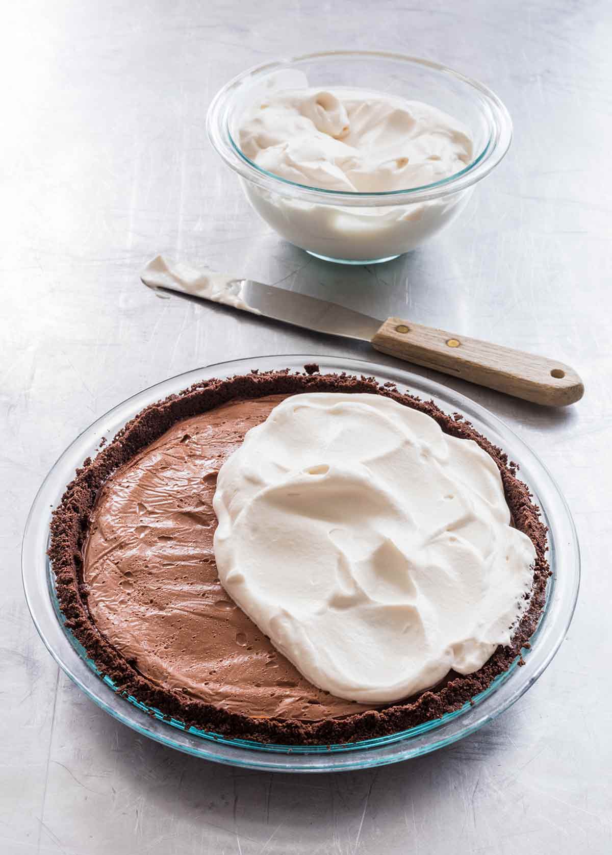 A dark chocolate cream pie partially covered with whipped cream and a knife and a bowl of whipped cream on the side.