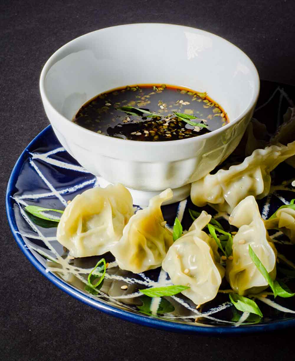 A bowl of dumpling dipping sauce on a blue and white plated with dumplings and sliced scallions.
