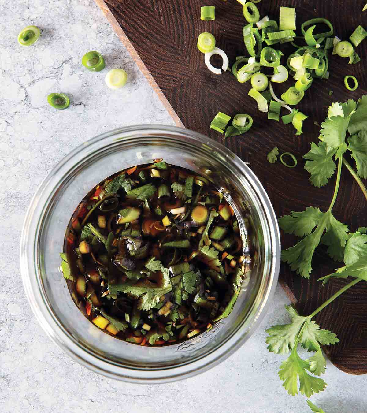 A glass bowl filled with dumpling dipping sauce with pieces of scallion and cilantro floating in it and scattered around the jar.