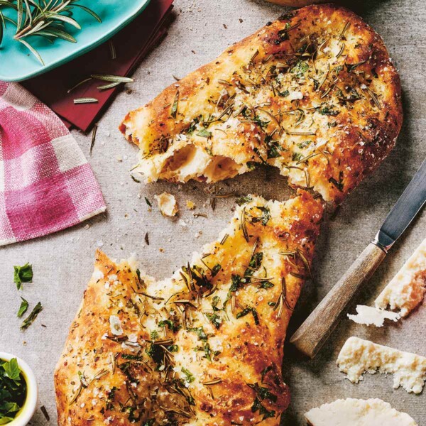 A torn loaf of focaccia with herbs and salt and sprigs of rosemary, a knife, and a hunk of pecorino on the side.