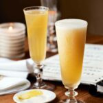 Two flutes filled with French 75 cocktail on a wooden table with a notebook and a plate of lemon slices nearby.