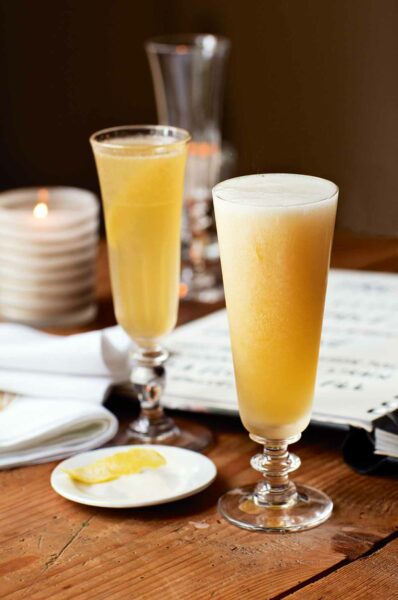 Two flutes filled with French 75 cocktail on a wooden table with a notebook and a plate of lemon slices nearby.