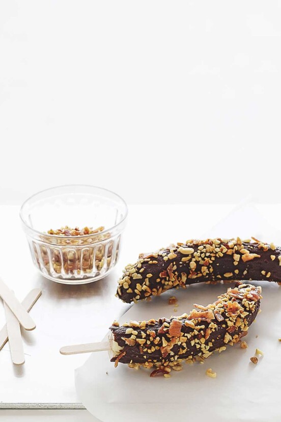 Two frozen chocolate-covered bananas on sticks with a bowl of peanuts and more popsicle sticks beside them.
