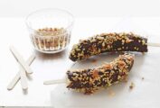Two frozen chocolate-covered bananas on sticks with a bowl of peanuts and more popsicle sticks beside them.