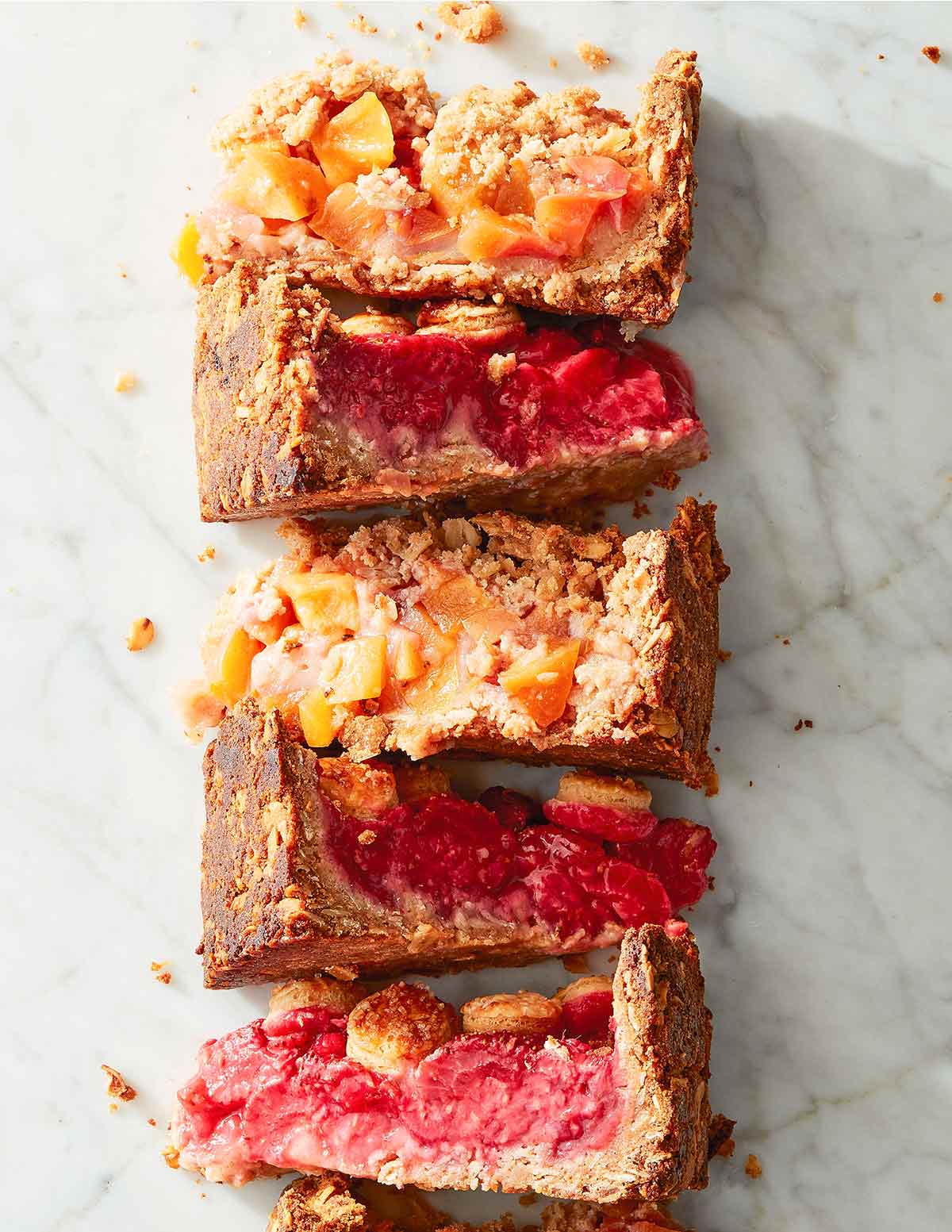 Pieces of fruit crostata with crumb topping, each filled with different fruit, lined up next to each other on a marble surface.