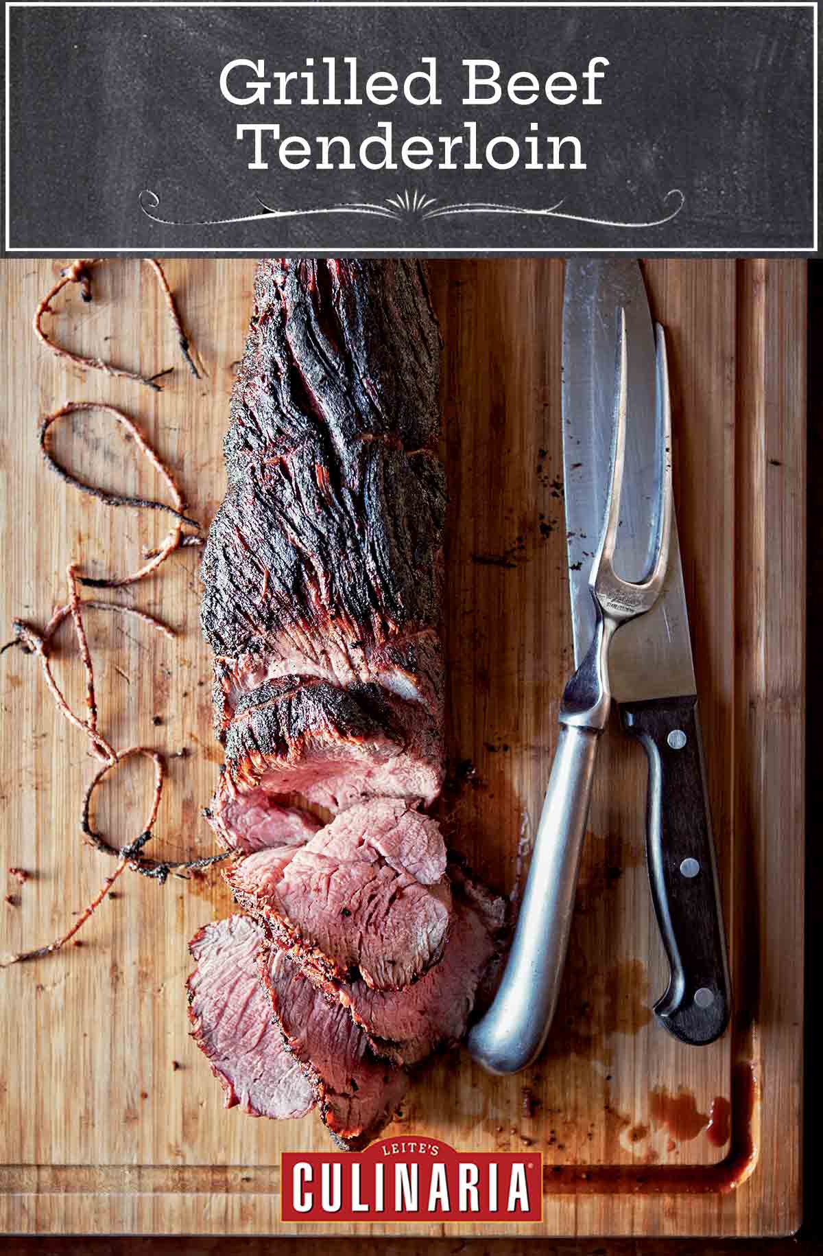 A whole grilled beef tenderloin, partially sliced on a wooden cutting board with a knife, meat fork, and twine beside it.