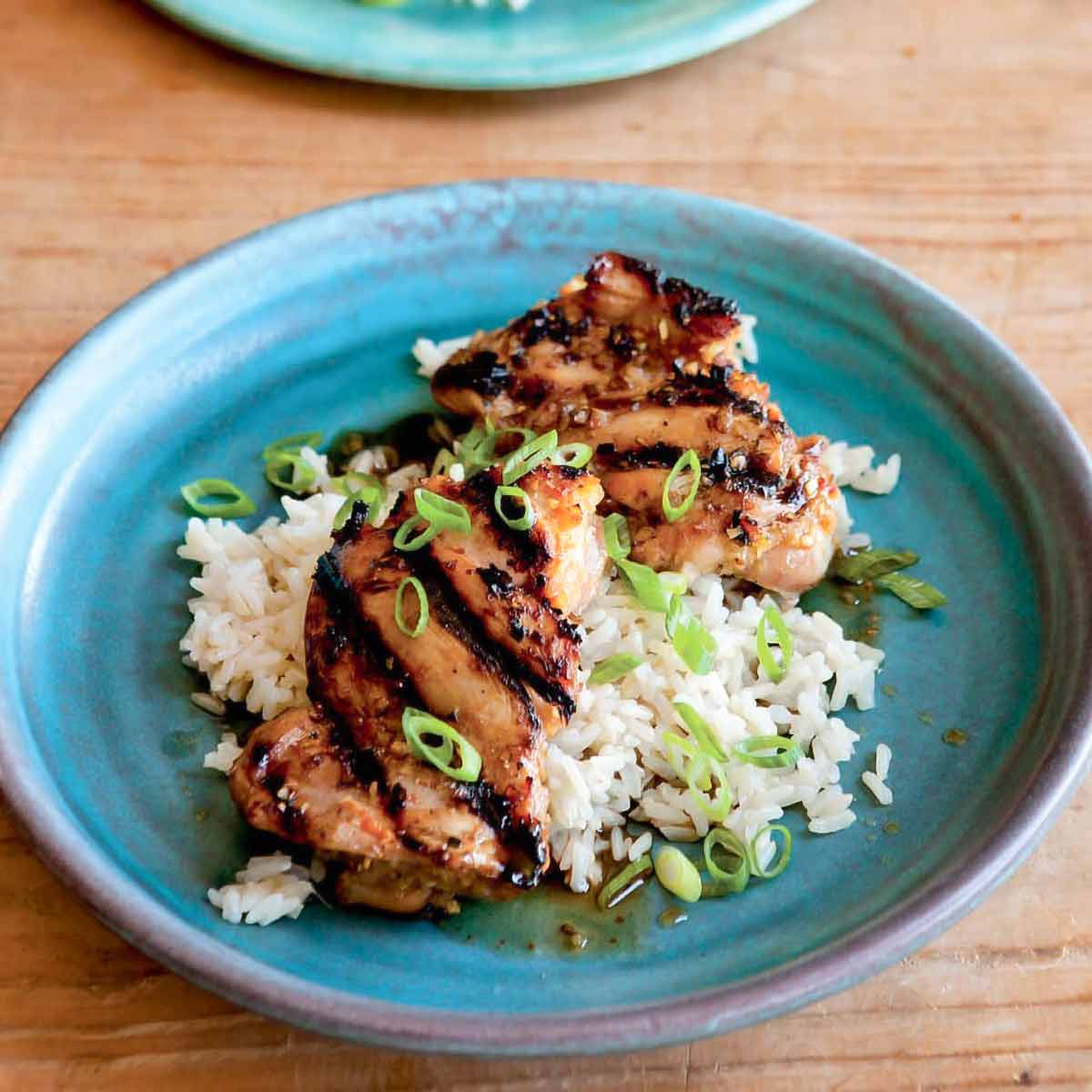 Two pieces of grilled lemongrass chicken on rice garnished with spring onions on a blue plate.