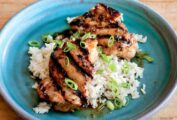 Two pieces of grilled lemongrass chicken on top of rice with scallion garnish on a blue plate.