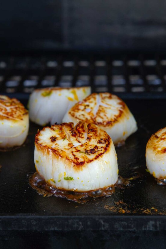 Several seared grilled scallops on a cast-iron griddle on a grill.