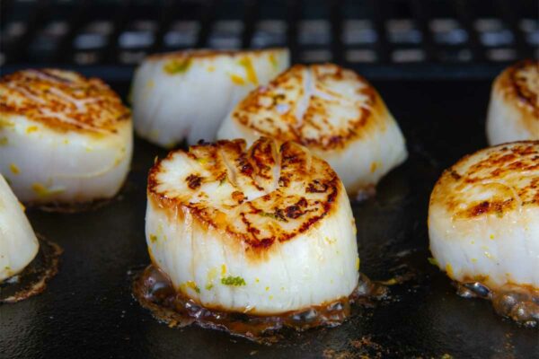 Several seared grilled scallops on a cast-iron griddle on a grill.