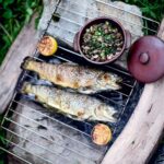 Two whole grilled trout on a grate over an ope fire with a dish of mushrooms and two grilled lemon halves on the side.