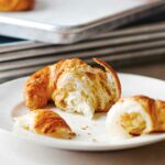 A torn croissant on a plate with more croissants on a rimmed baking sheet in the background.