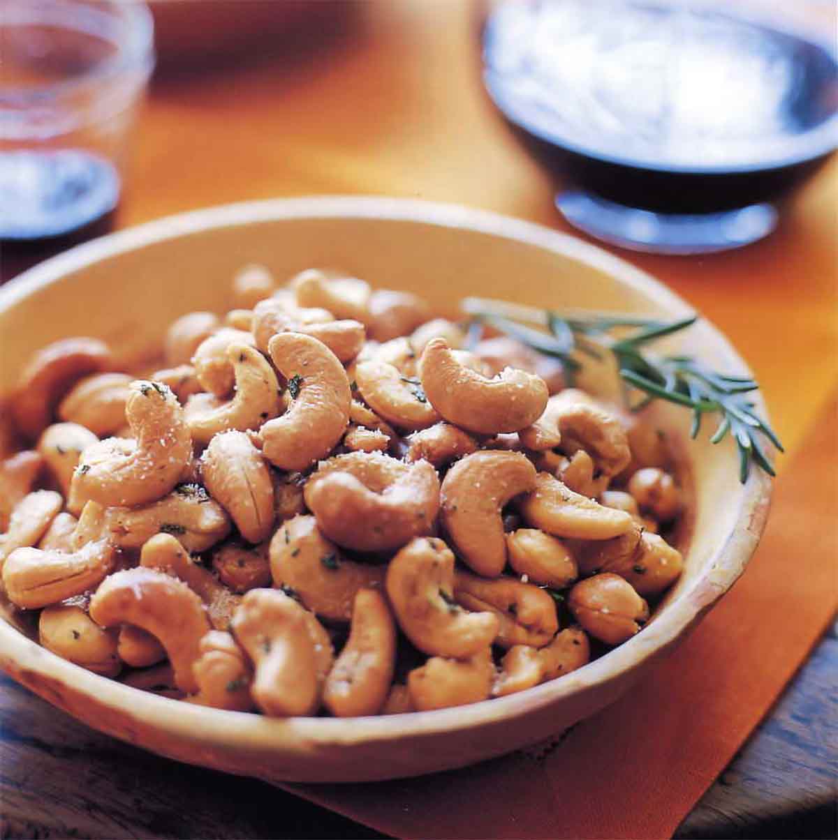 A bowl filled with Ina Garten's rosemary cashews with a sprig of rosemary on the side.