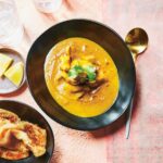 A black bowl filled with Macanese Portuguese chicken curry with dishes of naan and lemon wedges beside it.