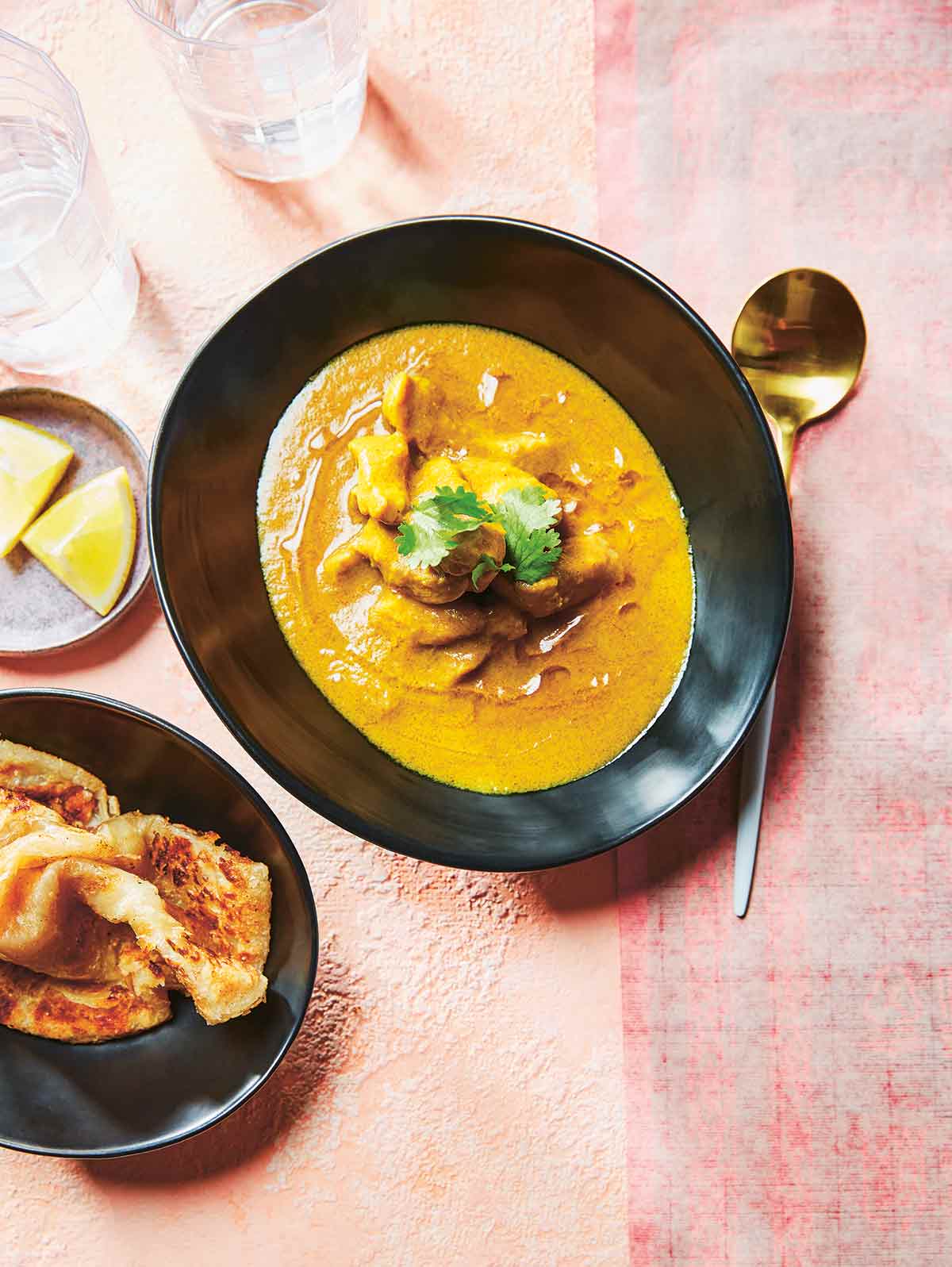 A black bowl filled with Macanese Portuguese chicken curry with dishes of naan and lemon wedges beside it.