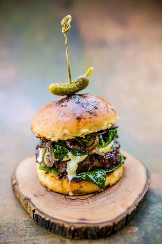 An example of how to make the best burger -- a burger piled with fixings and topped with a pickle on a wooden plate.