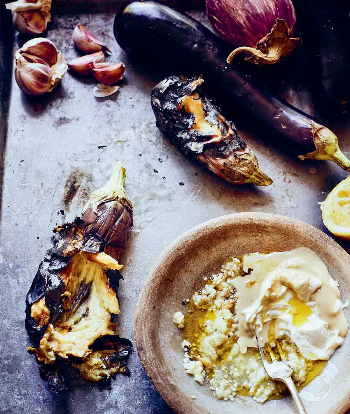 The components for moutabal -- eggplant, tahini, and yogurt -- being mixed in a bowl with a fork with a charred eggplant on the side.