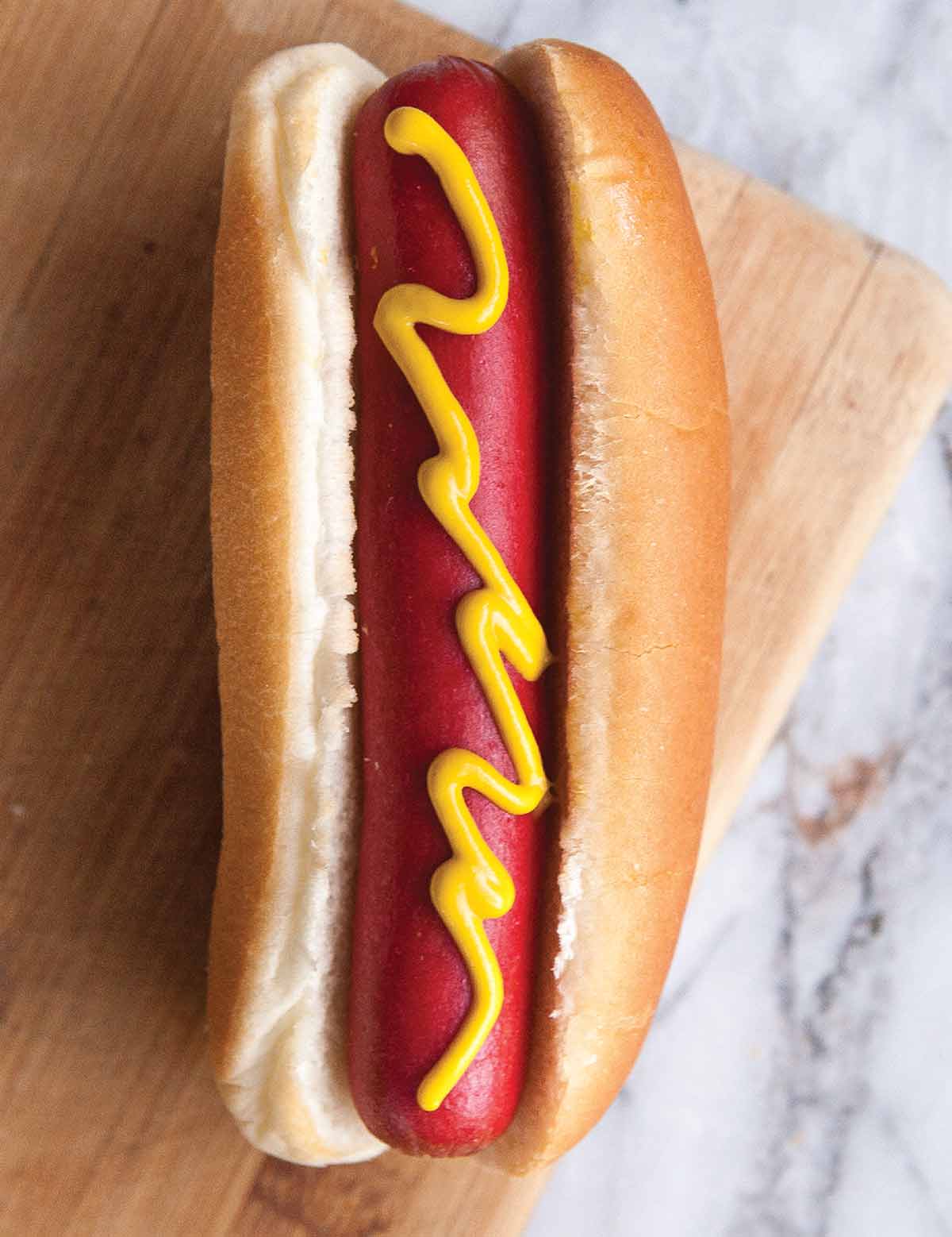 A perfect hot dog in a white bun, topped with a squiggle of mustard.